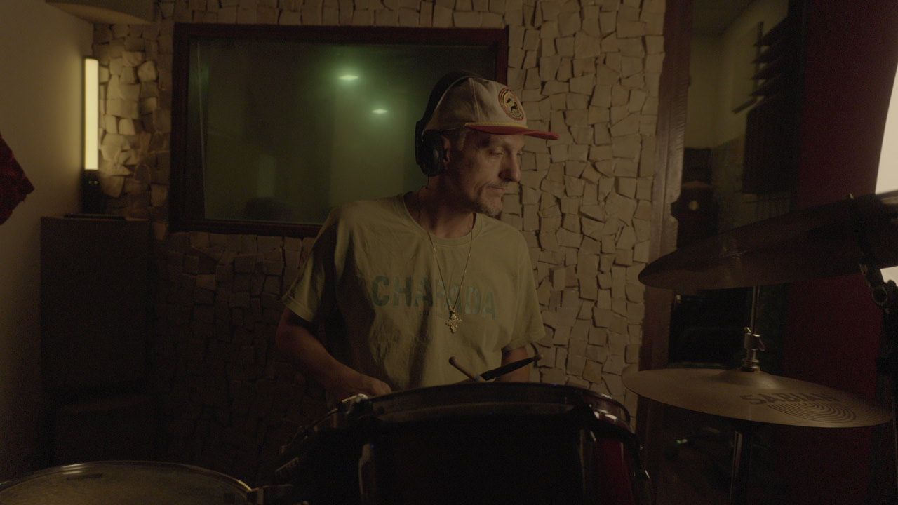 Gabriel Jahbê tocando bateria no estúdio com luz quente e parede de pedra ao fundo